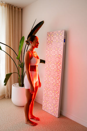 The Radiance960 full body red light panel used by a lady standing in bedroom with the LEDs glowing on the side of her body.