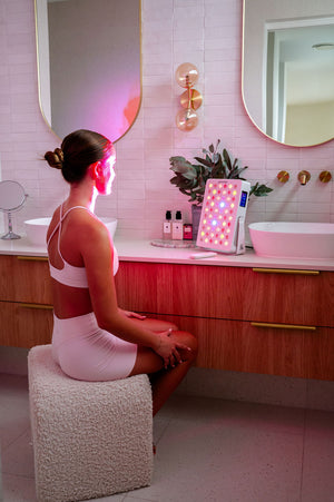 The Radiance60 red light panel used by a lady seated in the bathroom, panel placed on the benchtop, glowing LEDs on her body.